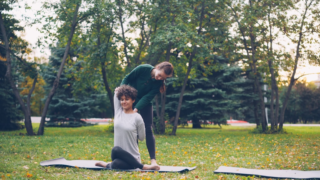 Diligent yoga student is learning Cow Face position sitting on mat and stretching arms while experienced teacher is helping her, girls are talking and laughing at outdoor ptactice.
