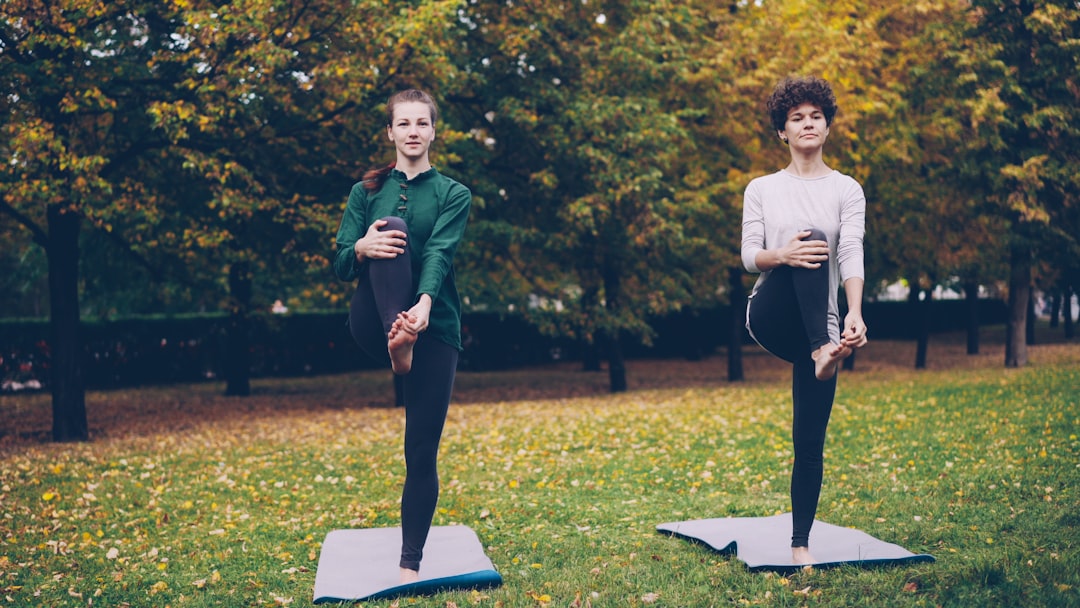 Couple of attractive girls are doing yoga in park practising balancing exercises standing on one leg during individual yoga practice with professional instructor.
